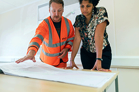 A woman and man review engieering plans spread over table top